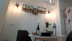 Ein Essbereich mit einem runden Tisch, Stühlen, einer Weinflasche, einer Wasserflasche und einer Vase mit Blumen. An der Wand sind Regale mit Pflanzen und eine Weltkarte angebracht.