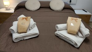 Ein ordentlich bezogenes Bett mit zwei Handtüchern, die mit einem 'Welcome'-Zettel verbunden sind, einer Nachttischlampe und einem Nachttisch mit Fernbedienung.