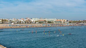 Ein Strand mit Menschen, die auf Paddleboards fahren und schwimmen, eine Steinmauer und ein Hotel im Hintergrund.