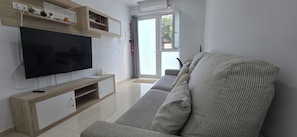 Un salon moderne doté d’un téléviseur à écran plat, d’un meuble de rangement en bois, d’un canapé gris et d’une porte-fenêtre coulissante donnant sur un balcon.