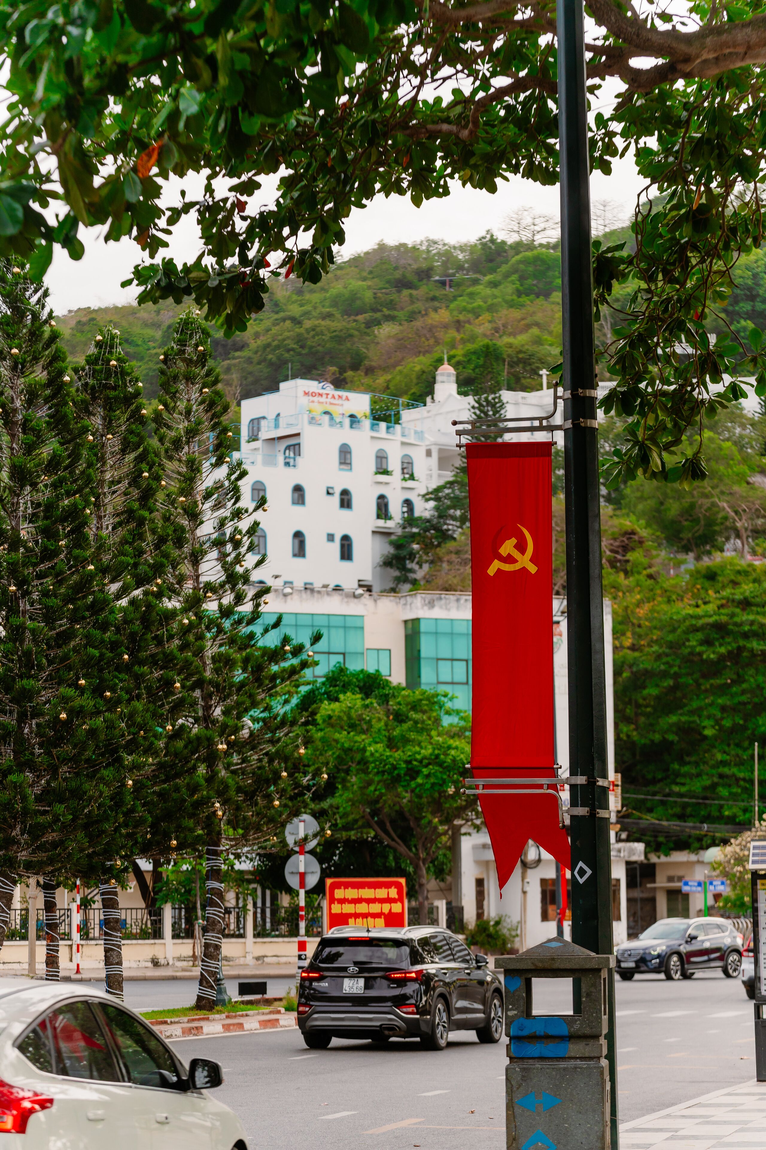 A street view with a red flag featuring a hammer and sickle emblem, a white building with "MONTANA" signage, and a black car in the foreground.