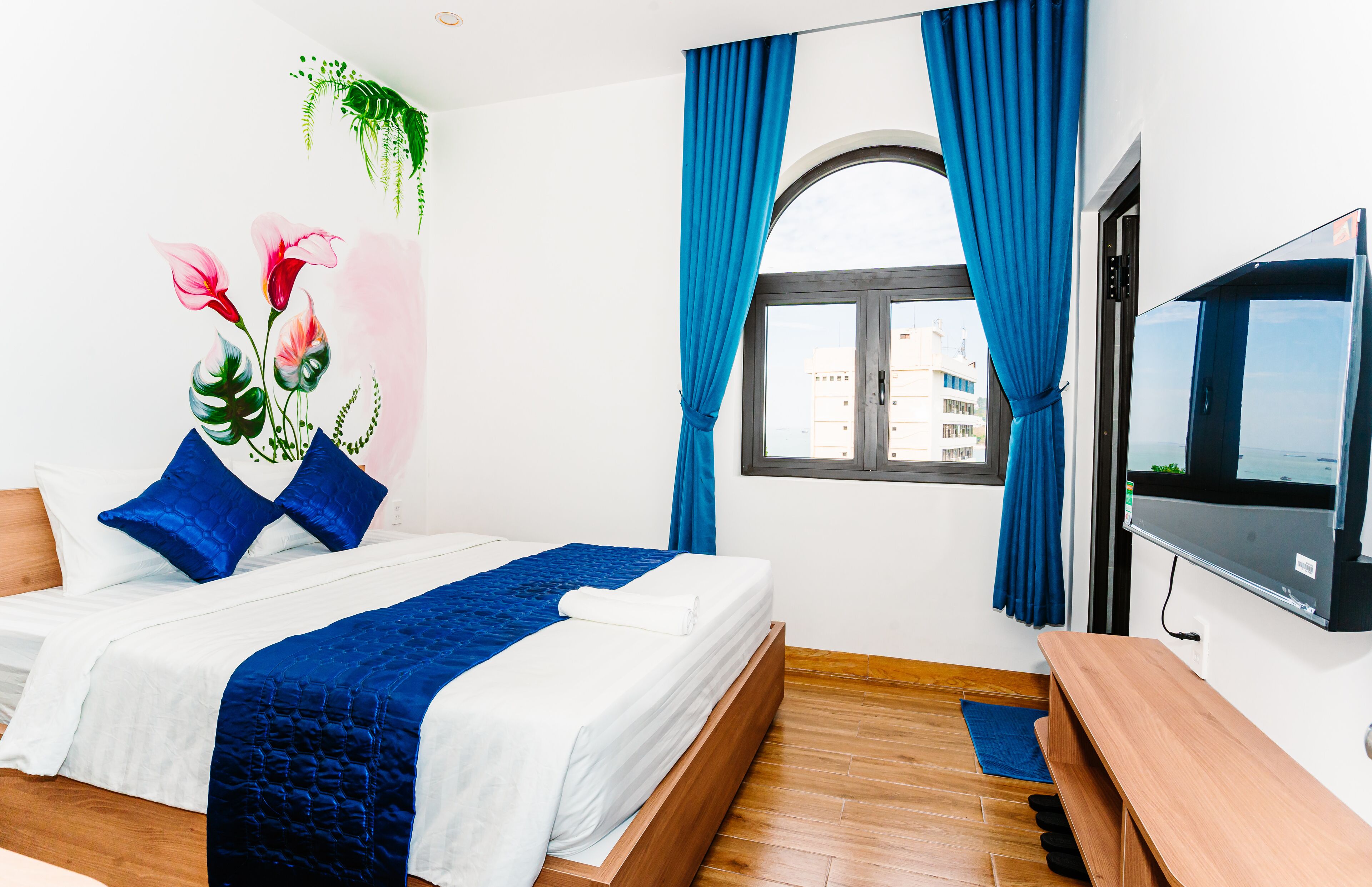 A hotel room with a bed, a TV, a window with curtains, and a view of buildings outside.