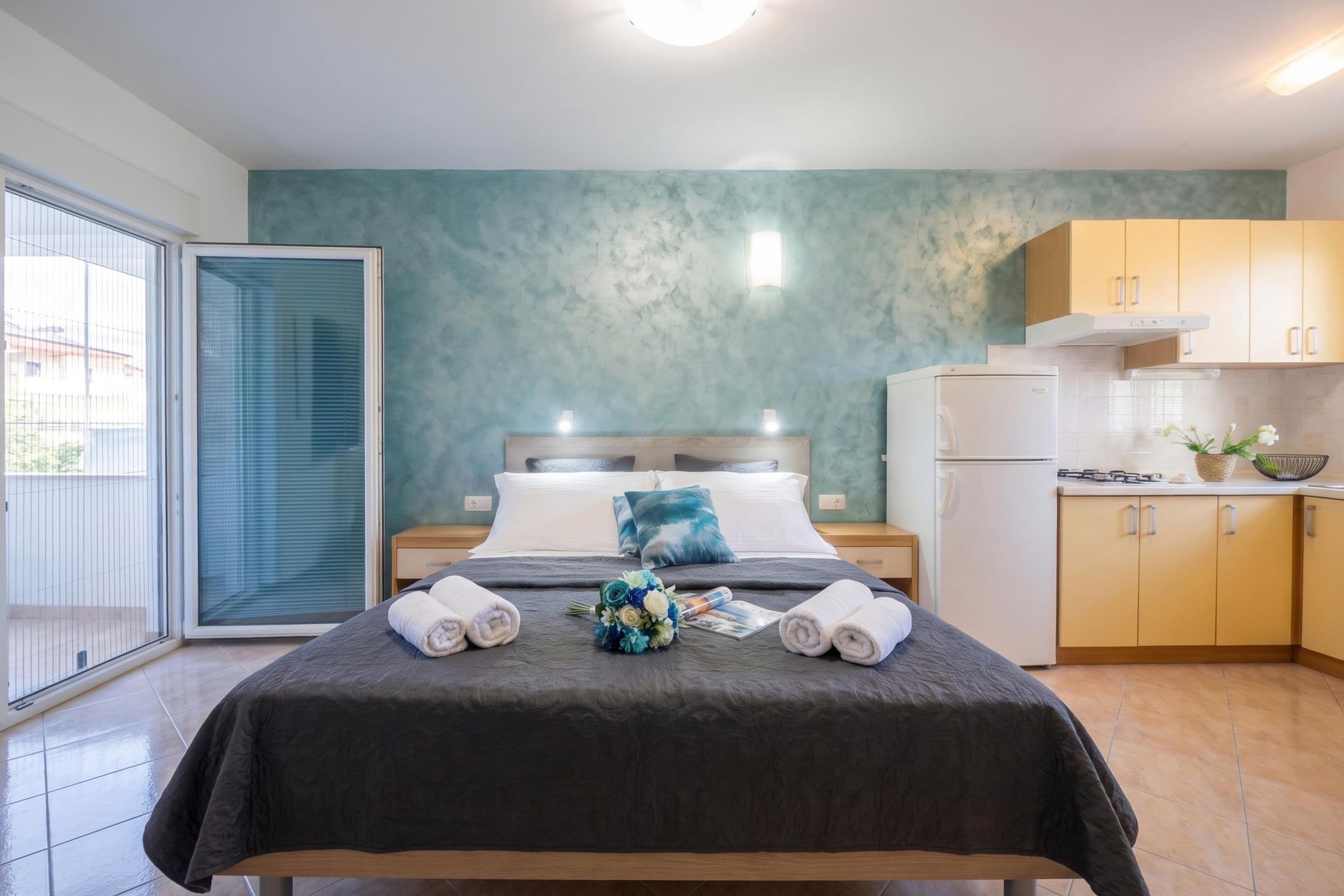 Ein Hotelzimmer mit Bett, einem kleinen Nachttisch, einem Bad mit Dusche, einer Küchenzeile mit Kühlschrank und Blick durch eine Schiebetür.