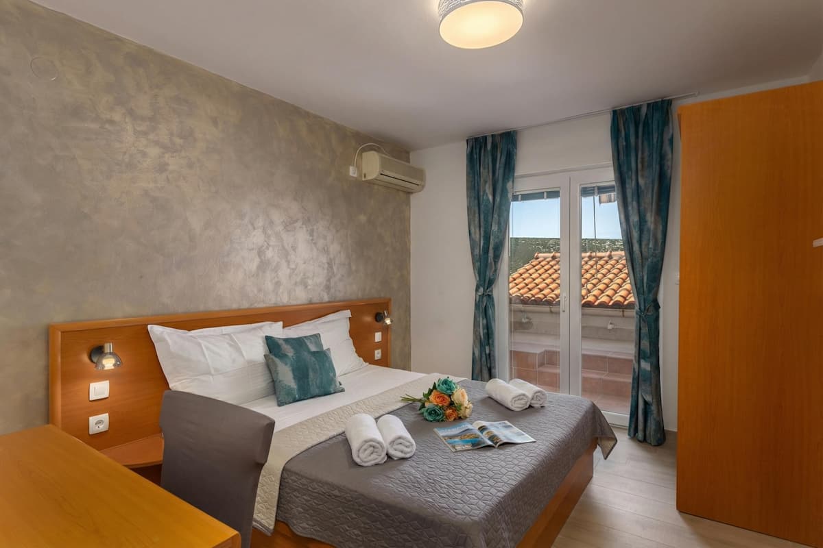 Une chambre d’hôtel comprenant un lit, une table de chevet en bois, une table, une fenêtre avec des rideaux et un appareil de climatisation.