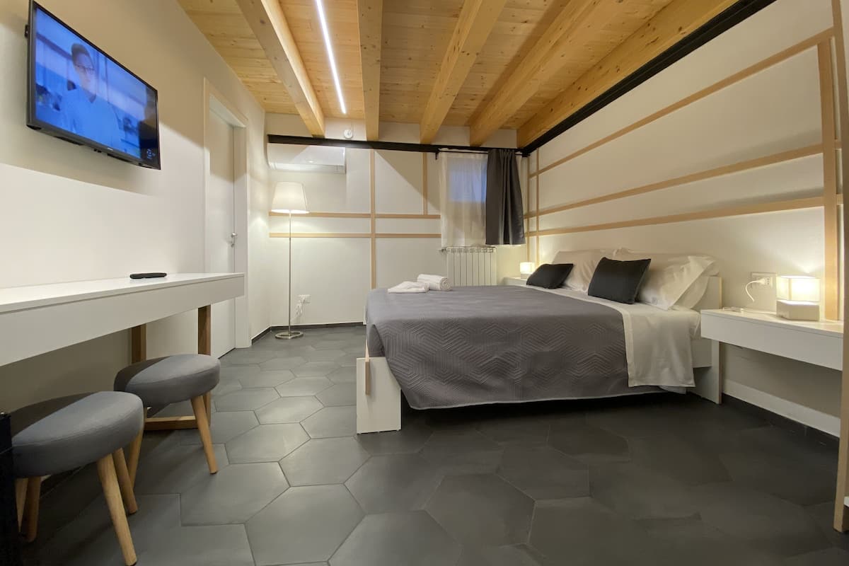 Une chambre d’hôtel moderne avec un plafond en bois, un lit recouvert d’une couverture grise, une table de chevet avec une lampe, un bureau avec une chaise et un téléviseur fixé au mur.
