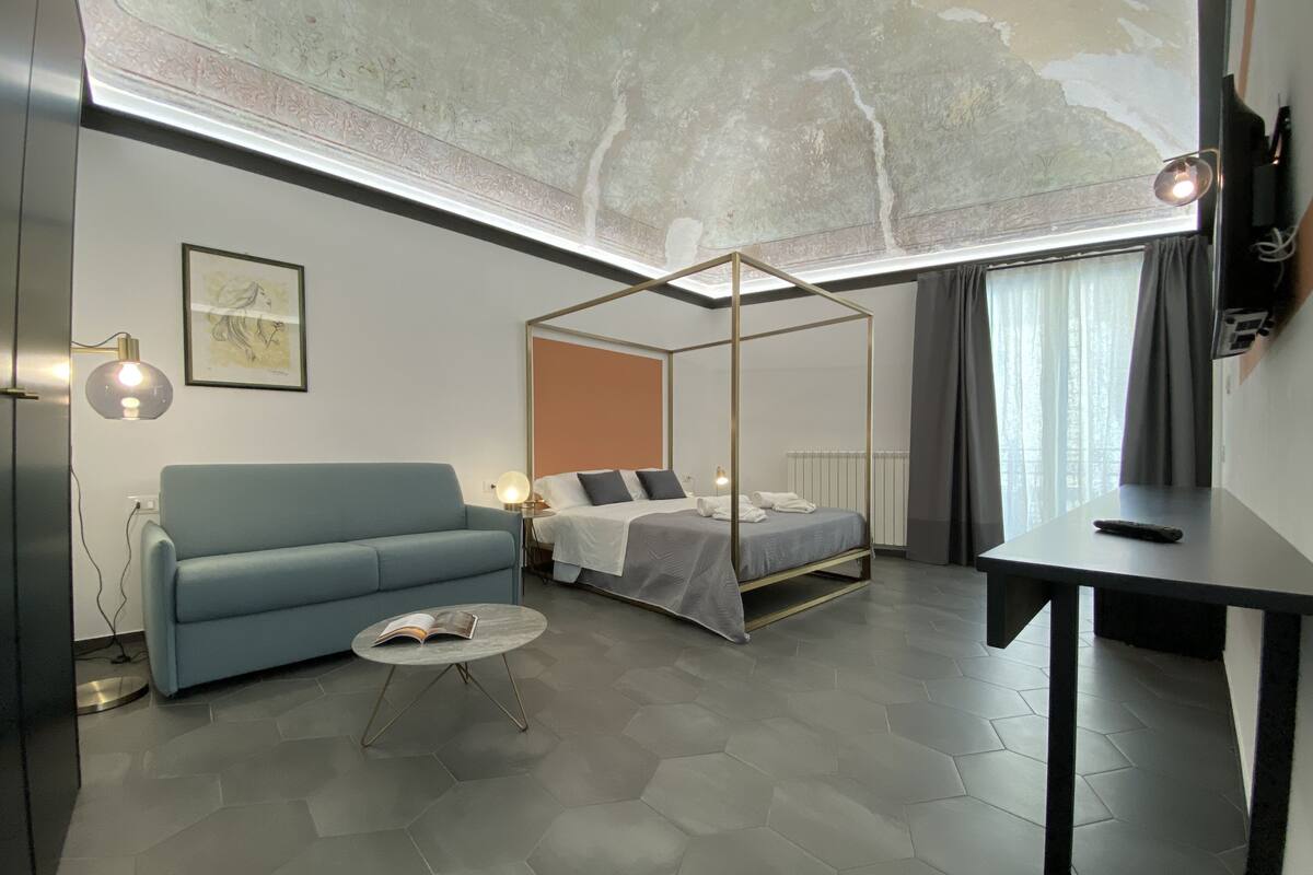 Une chambre d’hôtel moderne avec un lit à baldaquin, un canapé, une petite table ronde avec un livre et un bureau avec une télécommande.