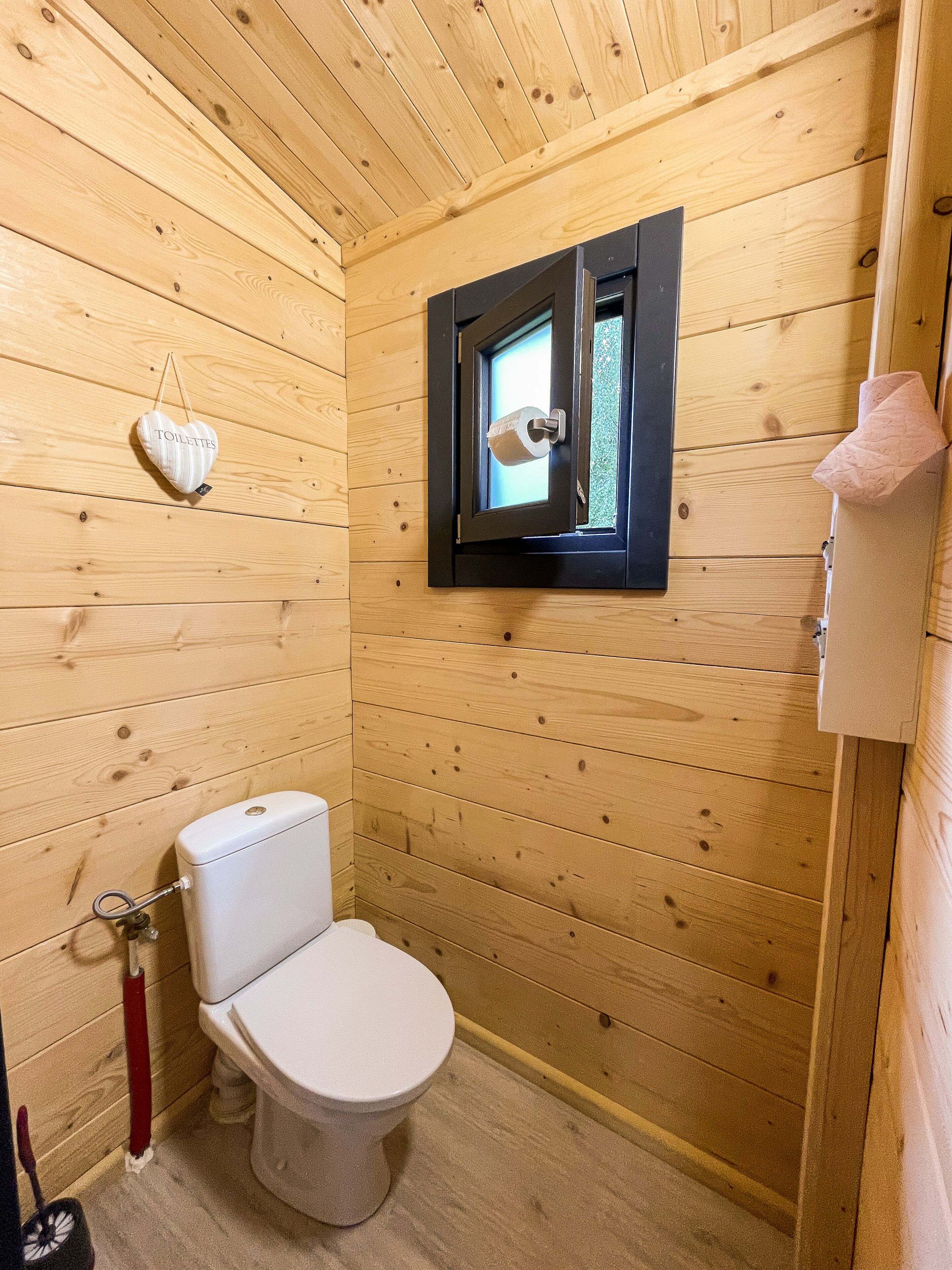 Ein kleines Badezimmer aus Holz mit Toilette, Fenster und einem Toilettenpapierhalter.