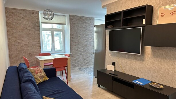 Ein modernes Wohnzimmer mit einem blauen Sofa, einem Esstisch mit roten Stühlen, einem an der Wand montierten Fernseher und einer Wand mit Muster.