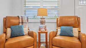 Dos sillones de cuero con cojines azules, una mesita de madera con una lámpara y libros, y una ventana con persianas en el fondo.