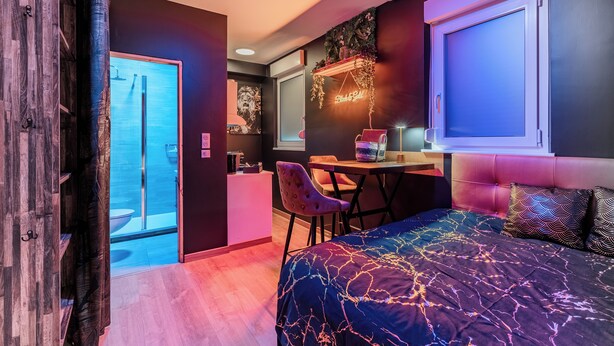 Un dormitorio moderno con una cama, un escritorio y una silla. Se ve un baño con ducha y retrete a través de una puerta abierta.