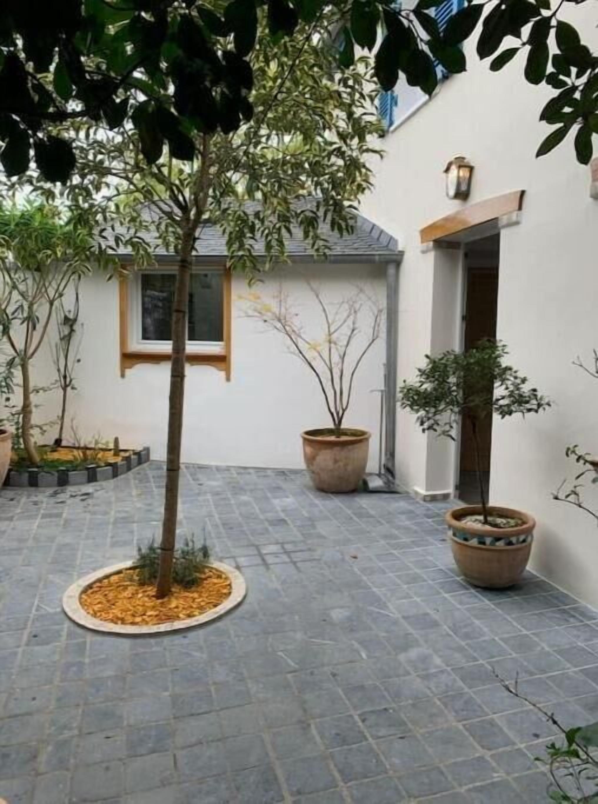 Une cour avec un sol carrelé, plusieurs plantes en pot et un bâtiment doté d’une fenêtre et d’une porte.