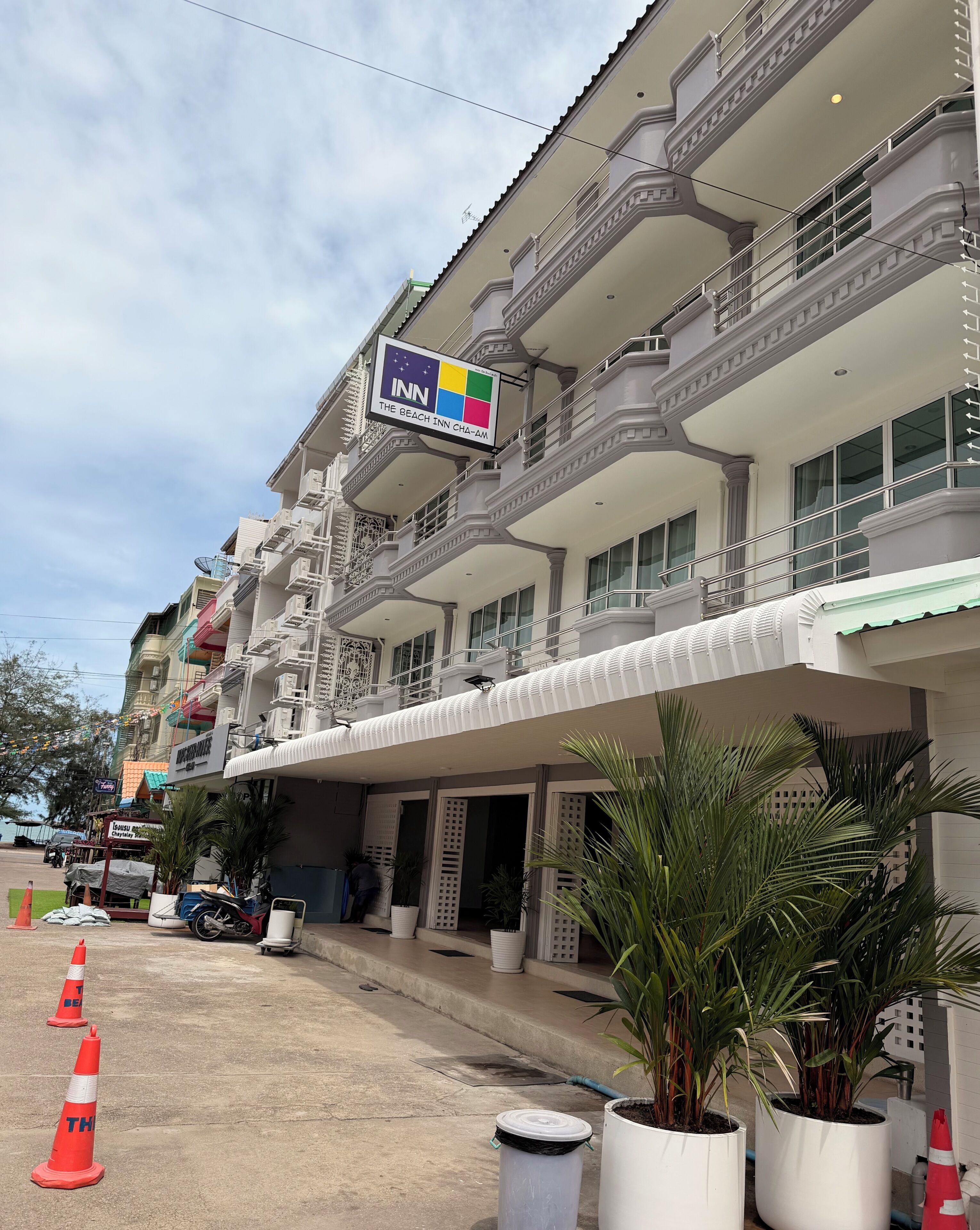 Un hotel de varios pisos con balcones, un letrero con temática playera y plantas en macetas.