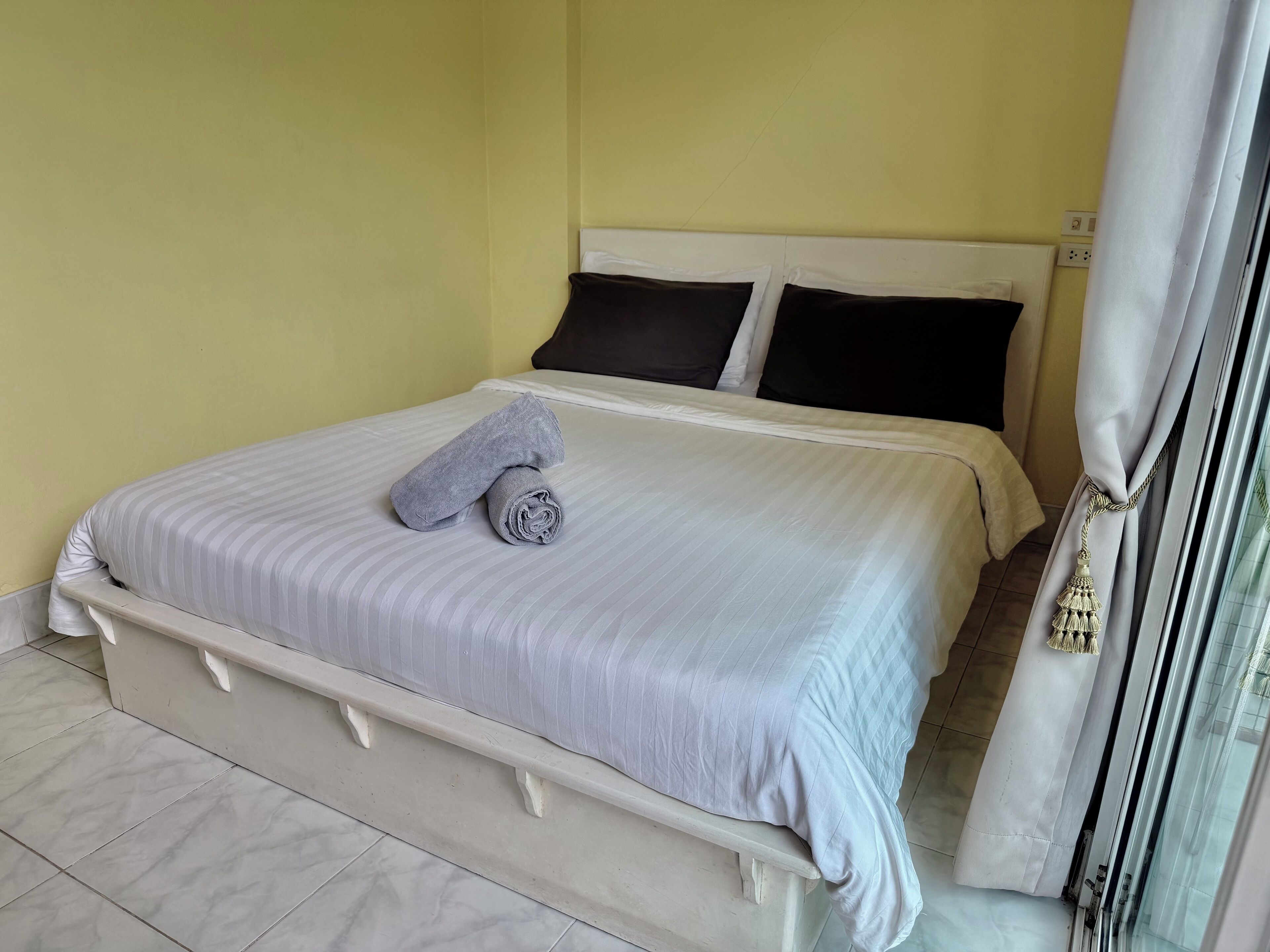 A neatly made bed with a rolled towel, a white headboard, and a window with a curtain.