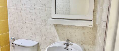 Une salle de bain avec des toilettes, un lavabo et un miroir. Les murs sont recouverts d’un papier peint à motifs.