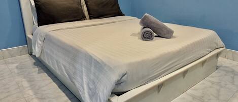 A neatly made bed with a white headboard and two dark pillows, a rolled towel on the footboard, and a blue wall in the background.