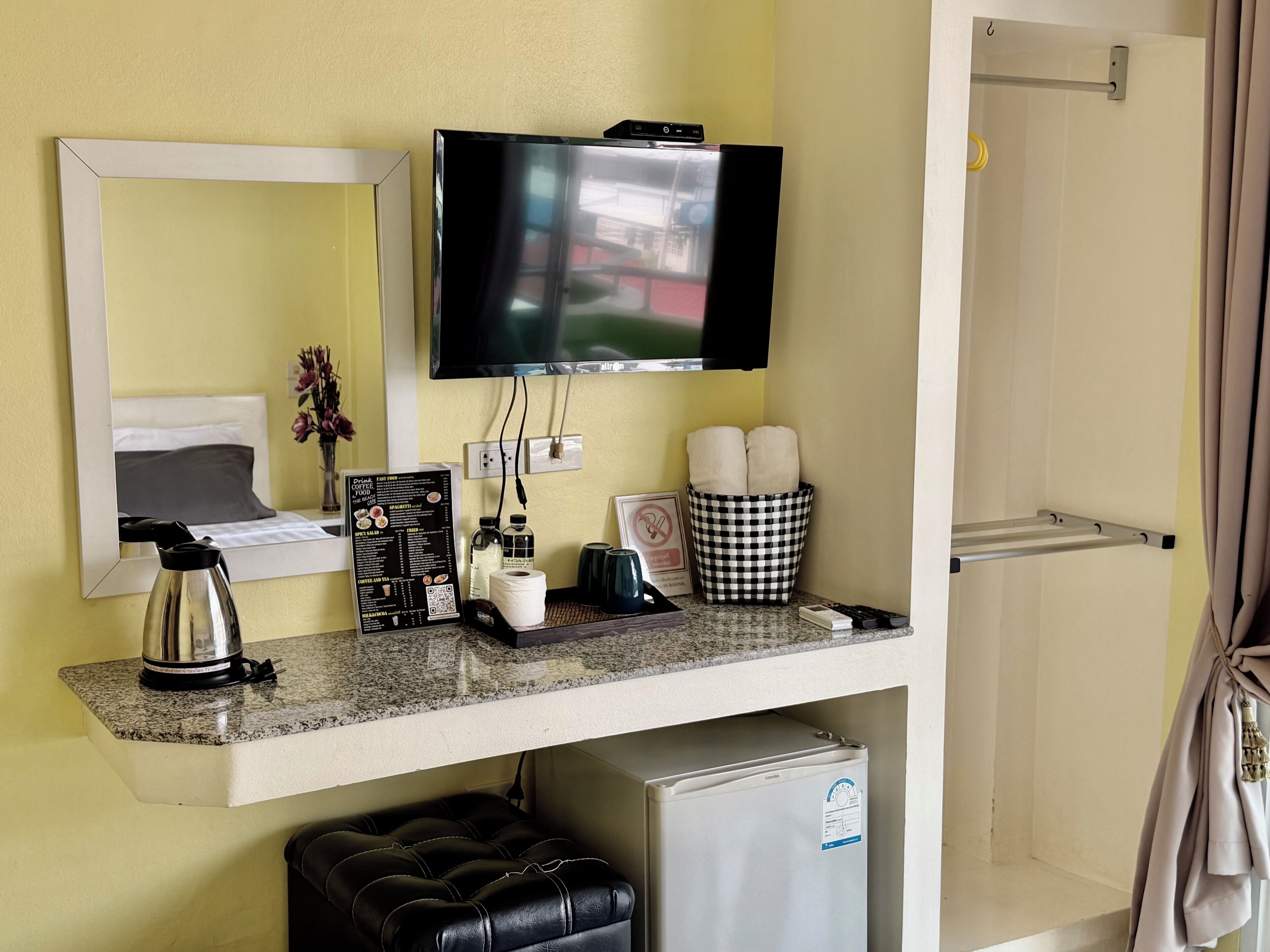 Une chambre d’hôtel équipée d’un téléviseur fixé au mur, d’un petit réfrigérateur, d’une machine à café, d’un panier à carreaux et d’un miroir qui reflète la pièce.