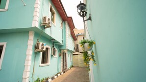 A narrow alleyway between buildings with a light blue facade, a black hanging lamp, and an air conditioning unit. - Royalty Place Hotel (Lagos)