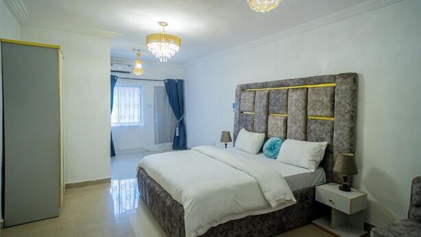A bedroom with a large bed, a nightstand, a lamp, and a chandelier. - Royalty Place Hotel (Lagos)