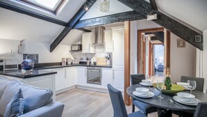 Una cocina moderna con armarios blancos, un comedor con mesa redonda para seis comensales y un dormitorio en entrepiso con vigas a la vista.