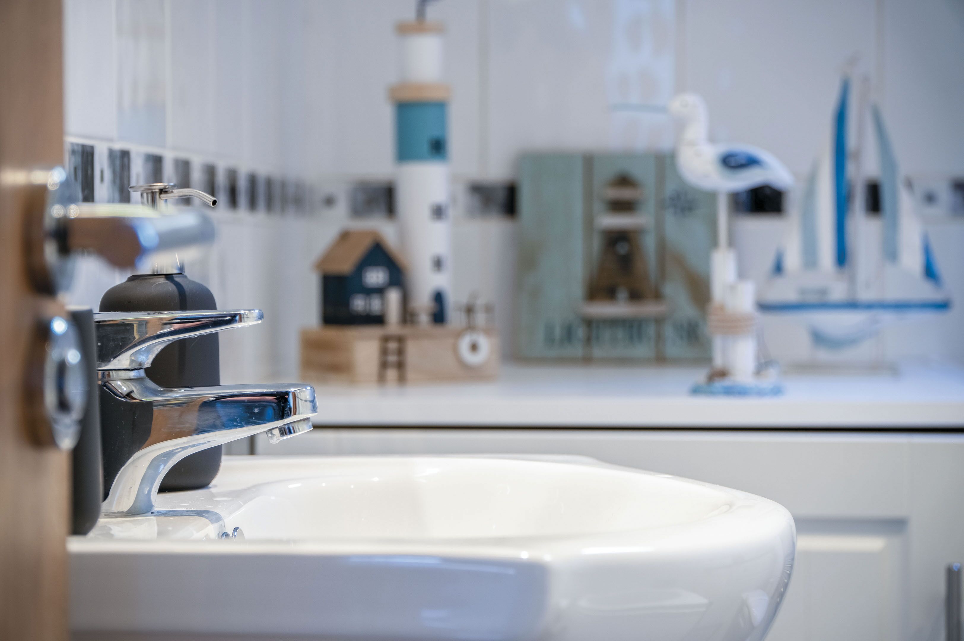 Une salle de bain avec un lavabo blanc, un robinet chromé et un miroir qui reflète un modèle de phare, un petit modèle de bateau et un meuble.