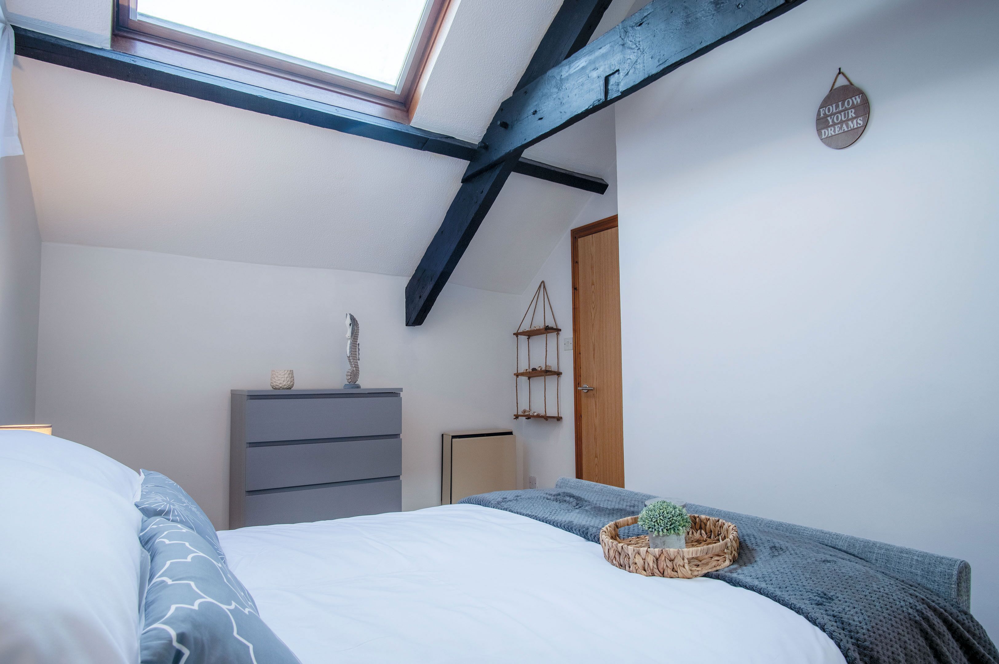 Un dormitorio con techo inclinado, una cama con manta gris, un aparador, una planta pequeña y un letrero motivacional en la pared.