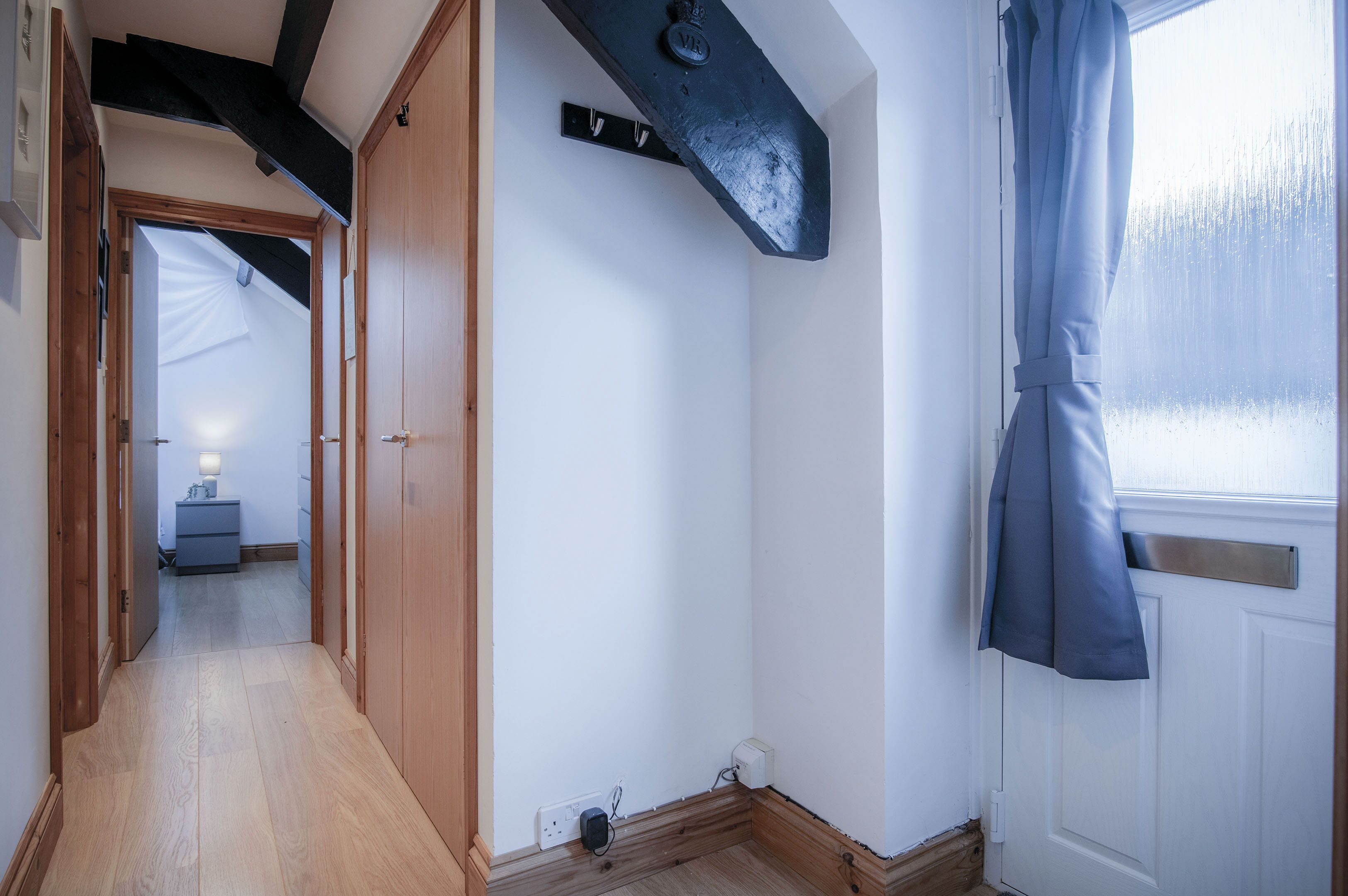 A narrow hallway with wooden flooring, a white door, and a window with a blue curtain.