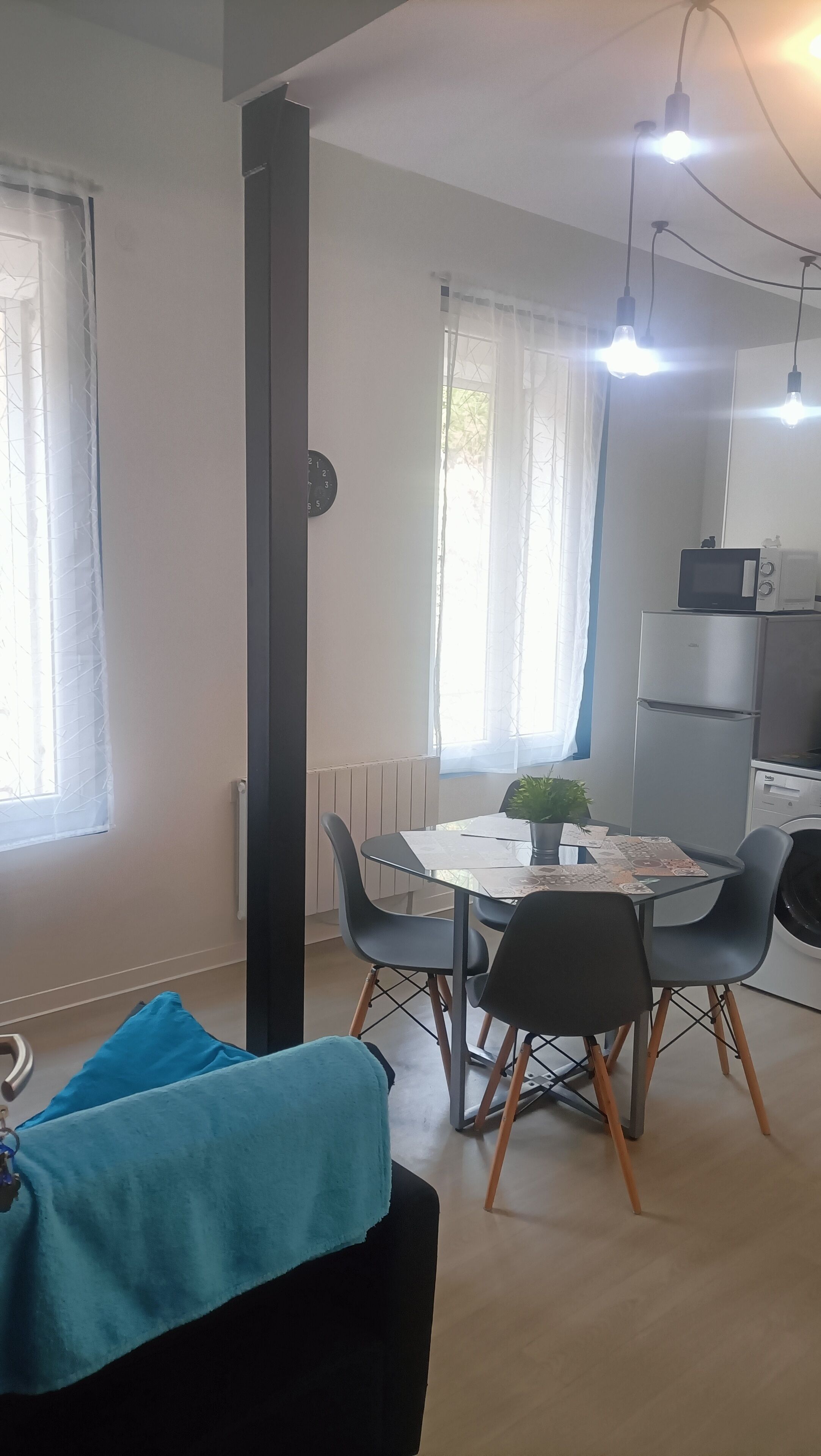 Uma cozinha moderna com frigorífico, micro-ondas e pia. Há uma mesa de jantar com cadeiras e uma planta em vaso. O espaço possui grandes janelas com cortinas leves.