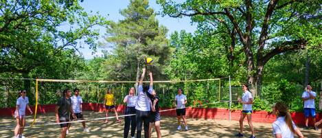 Un grupo de personas juega voleibol en una cancha al aire libre rodeada de árboles.