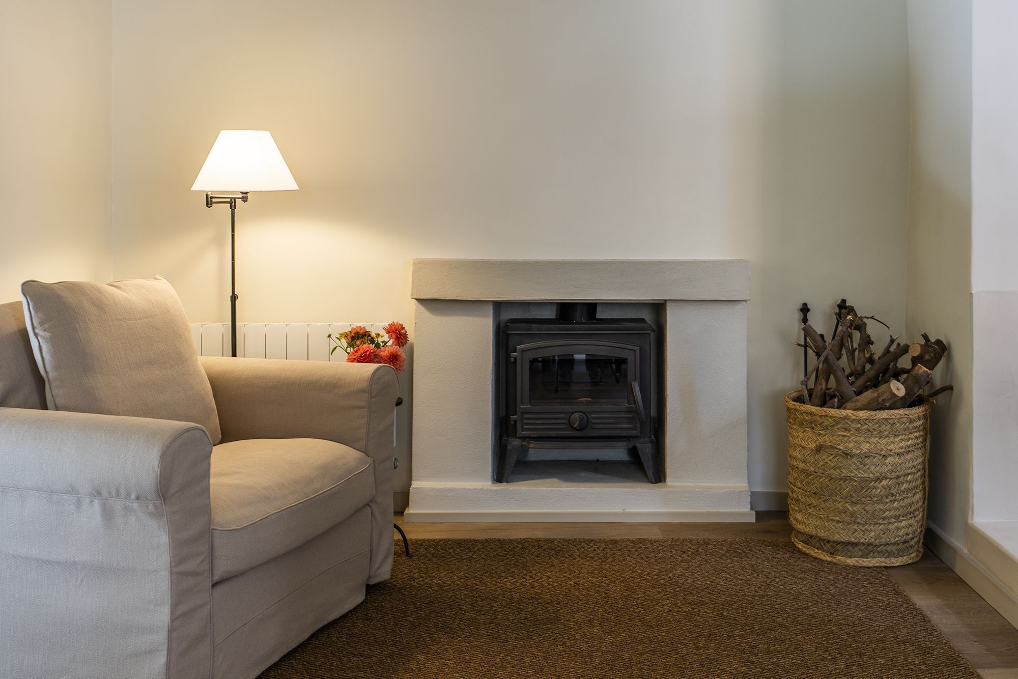 A living room with a beige sofa, a floor lamp, a fireplace, and a woven basket with logs.