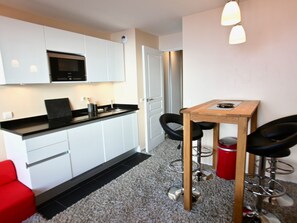 A compact kitchen with white cabinets, a black countertop, and a microwave. There is a small dining area with a wooden table and two black chairs. A red trash bin is visible. - Cozy 2-room apartment for 4, Southeast balcony, ski-in/ski-out in Avoriaz (Avoriaz)