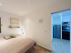 Un dormitorio con cama, lámpara de noche, un cuadro en la pared y puerta que da a una zona de cocina con refrigerador y horno.