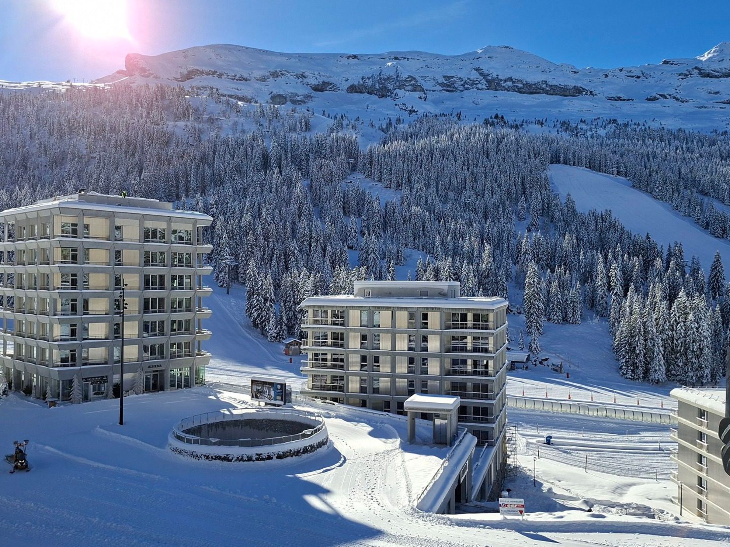 A snow-covered mountain resort with modern buildings, a ski lift, and a clear view of the surrounding forest.