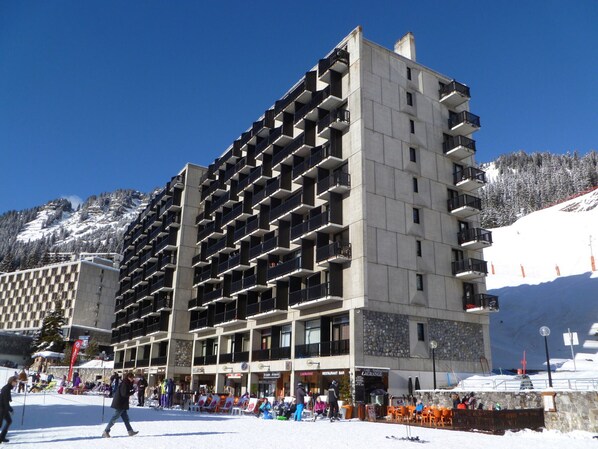 Hotel com vários andares e varandas, com uma estância de esqui ao fundo e áreas de estar ao ar livre.