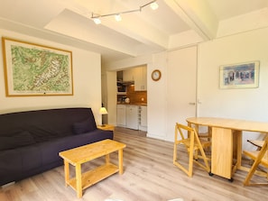 A living room with a dark sofa, a wooden coffee table, a dining table with chairs, a kitchen in the background, and artwork on the walls.