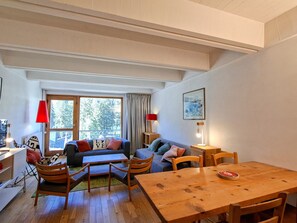 A modern living room with a sofa, chairs, a wooden table, and a large window with curtains.