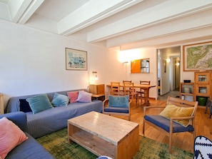 Ein Wohnzimmer mit einer Couch, einem Couchtisch und Sesseln. Ein Esstisch mit Stühlen und eine Lampe sind ebenfalls vorhanden. An der Wand hängt ein Gemälde, und zur Rechten eine Karte.