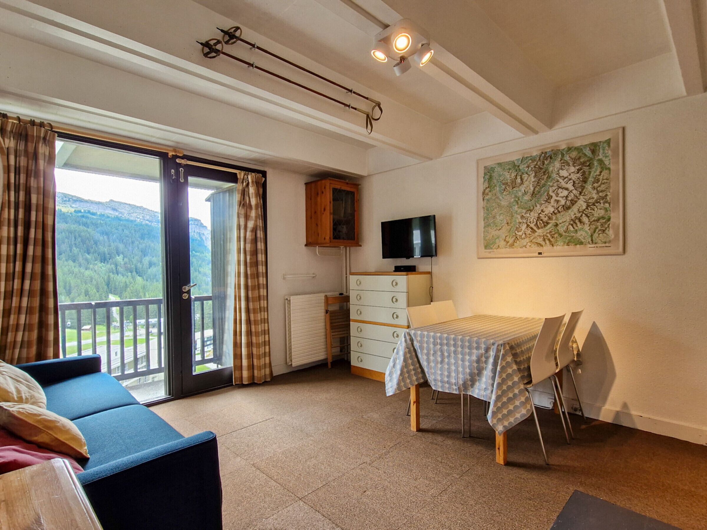 Ein Zimmer mit einer Couch, einem Esstisch mit Stühlen, einem Fernseher, einem Schrank und einem großen Fenster mit Vorhängen.