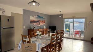 Comedor con una mesa preparada para cuatro, refrigerador, televisor y vista al exterior a través de una puerta corrediza de vidrio.