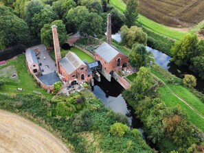 Luchtbeeld van een historisch industrieel gebouw met twee schoorstenen, een kanaal en omliggend groen.