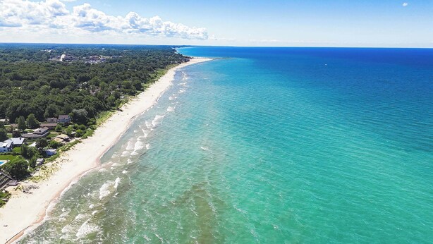Luftaufnahme einer Küstenlandschaft mit Sandstrand, klarem türkisblauem Wasser und einem dichten Waldgebiet mit Gebäuden.