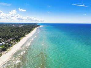 Vista aérea de una zona costera con playa arenosa, aguas turquesas cristalinas y un área boscosa densa con edificios.