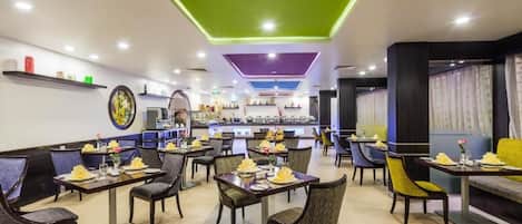 A restaurant with multiple dining tables and chairs, a bar area in the background, and a ceiling with green lighting accents.