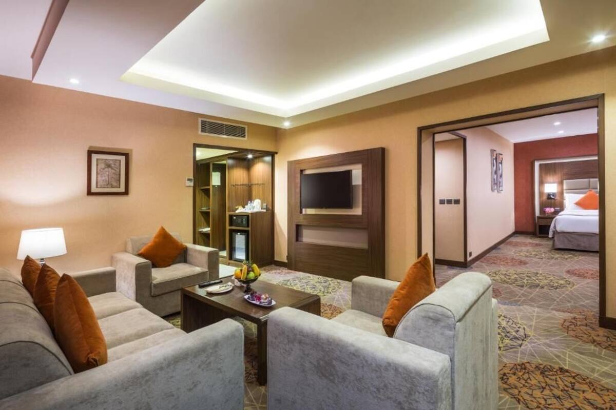 A hotel room with a sofa set, a television, a small table with fruit, and a bed with a nightstand.