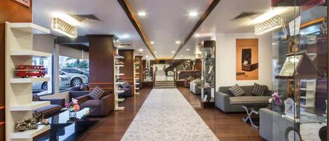 Un moderno vestíbulo de hotel con una escalera central, zonas de descanso y una exhibición de diversos objetos.