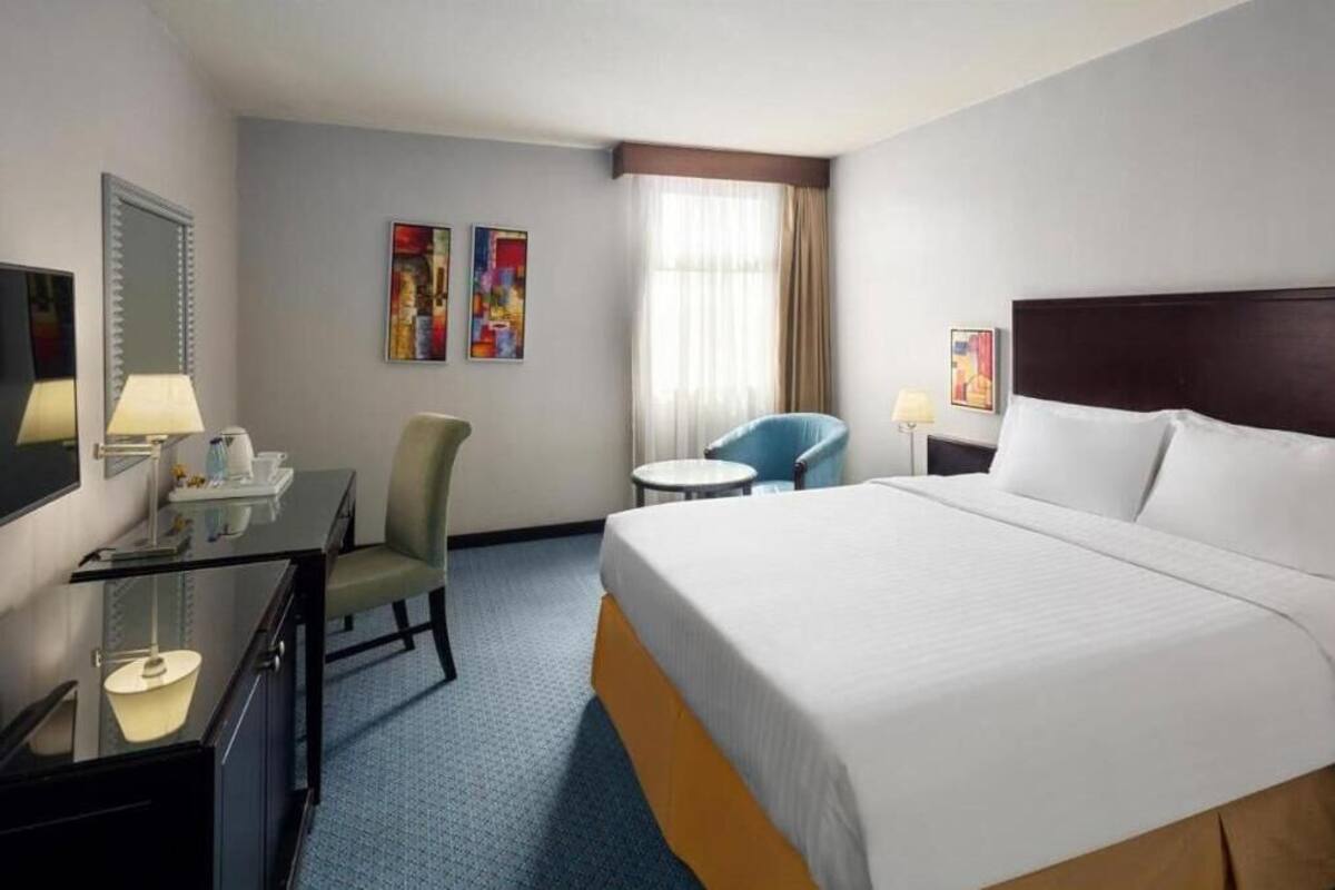 A hotel room with a large bed, a desk with a lamp, a chair, and two paintings on the wall.