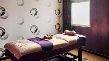 Un lit simple avec une couverture violette et une serviette, une petite boîte en bois et une fenêtre avec des rideaux.