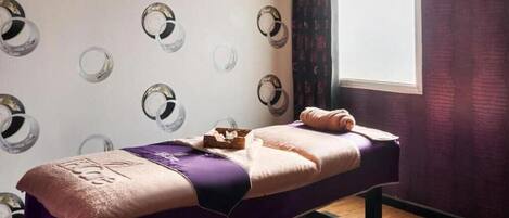 A single bed with a purple blanket and a towel, a small wooden box, and a window with curtains.