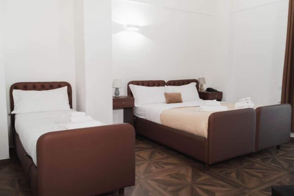 A hotel room with a double bed, a single bed, a nightstand with a lamp, and a brown patterned floor.