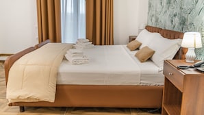 Una cama bien hecha con una manta color beige y almohadas, una mesita de noche de madera con una lámpara y un teléfono, y una pared con estampado de hojas tropicales.