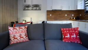 Una sala moderna con un sofá gris, dos cojines blancos y rojos con estampado, una pared de madera, un mueble blanco y una zona de cocina al fondo.